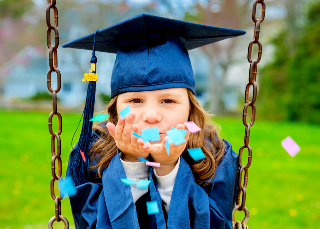 Kindergarten girl in graduation cap & gown blowing confetti